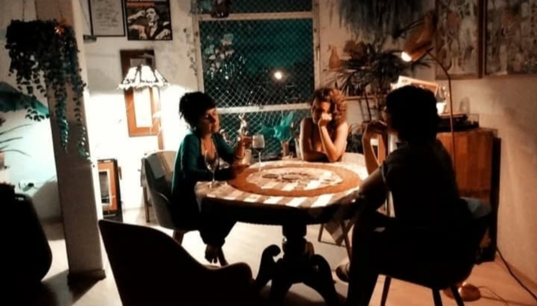 três mulheres sentadas em volta de uma mesa de madeira redonda. É um encontro que se passa a noite, em um ambiente cheio de plantas e quadros, possivelmente um vinho na mão de uma delas.