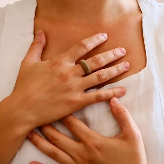 foto das mãos de uma mulher sobre o centro do peito. Há um anel de madeira na mão direita. Não é possível ver a cabeça ou os ombros, apenas as mãos sobre o centro do peito.